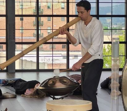 Musician plays a long wooden didgeridoo and taps a handpan and gong during a sound-healing session in a bright urban loft studio with floor-to-ceiling windows and people resting on mats.