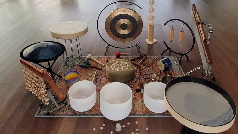 Zen-ready sound bath setup on hardwood floor: three white crystal singing bowls, brass gong, steel handpan, frame drum, chimes, mallets and a meditation cushion arranged on a decorative rug with crystals — wellness/meditation studio instruments.