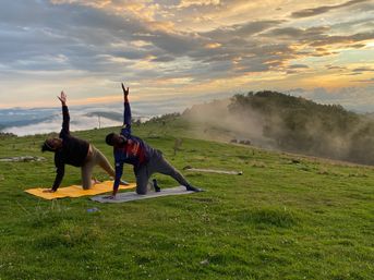 Two people doing yoga on mats in a misty mountain meadow at sunrise, stretching toward the sky with rolling green hills, grazing animals and dramatic cloud-filled sky in the background