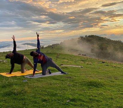 Two people doing yoga on mats in a misty mountain meadow at sunrise, stretching toward the sky with rolling green hills, grazing animals and dramatic cloud-filled sky in the background