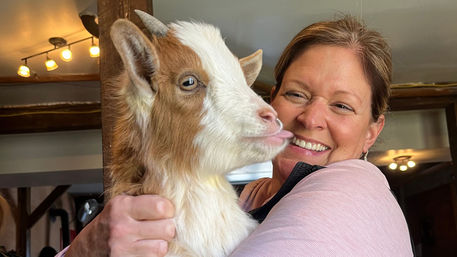 Smiling woman holding a brown-and-white baby goat with its tongue out inside a cozy farmhouse room with exposed beams and warm lighting