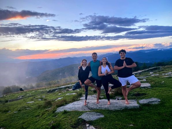 Four barefoot people balancing in tree pose on a rocky mountain ridge at sunset, colorful clouds and layered blue mountains stretching into the distance.