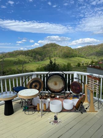 Sunlit wooden deck arranged with gongs, drums, crystal singing bowls and chimes overlooking lush rolling green hills and a bright blue sky with fluffy clouds.