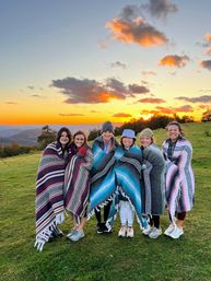 Six friends wrapped in colorful striped blankets, smiling on a grassy mountain meadow at sunset with glowing clouds and distant ridgelines.