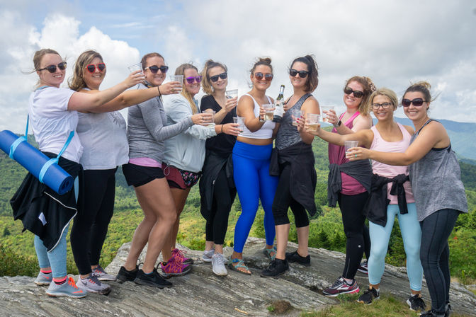 Group of women friends toasting with drinks on a rocky mountain overlook, wearing activewear and sunglasses with hiking gear and a yoga mat visible, green hills and cloudy sky in the background.