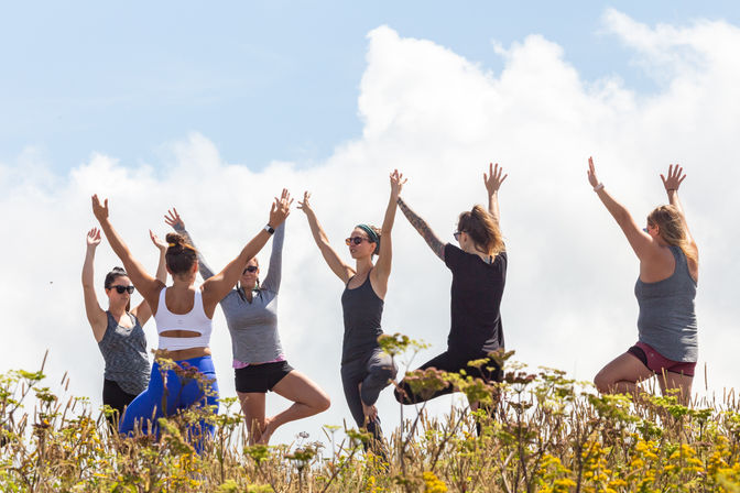 Group of women in tree pose practicing outdoor yoga on a wildflower-covered hilltop beneath a bright, cloudy sky