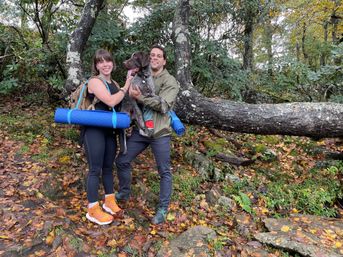 Two hikers on a leafy woodland trail holding a happy brown-and-white hunting dog beside a mossy fallen log, both wearing backpacks with blue rolled yoga mats amid autumn leaves.