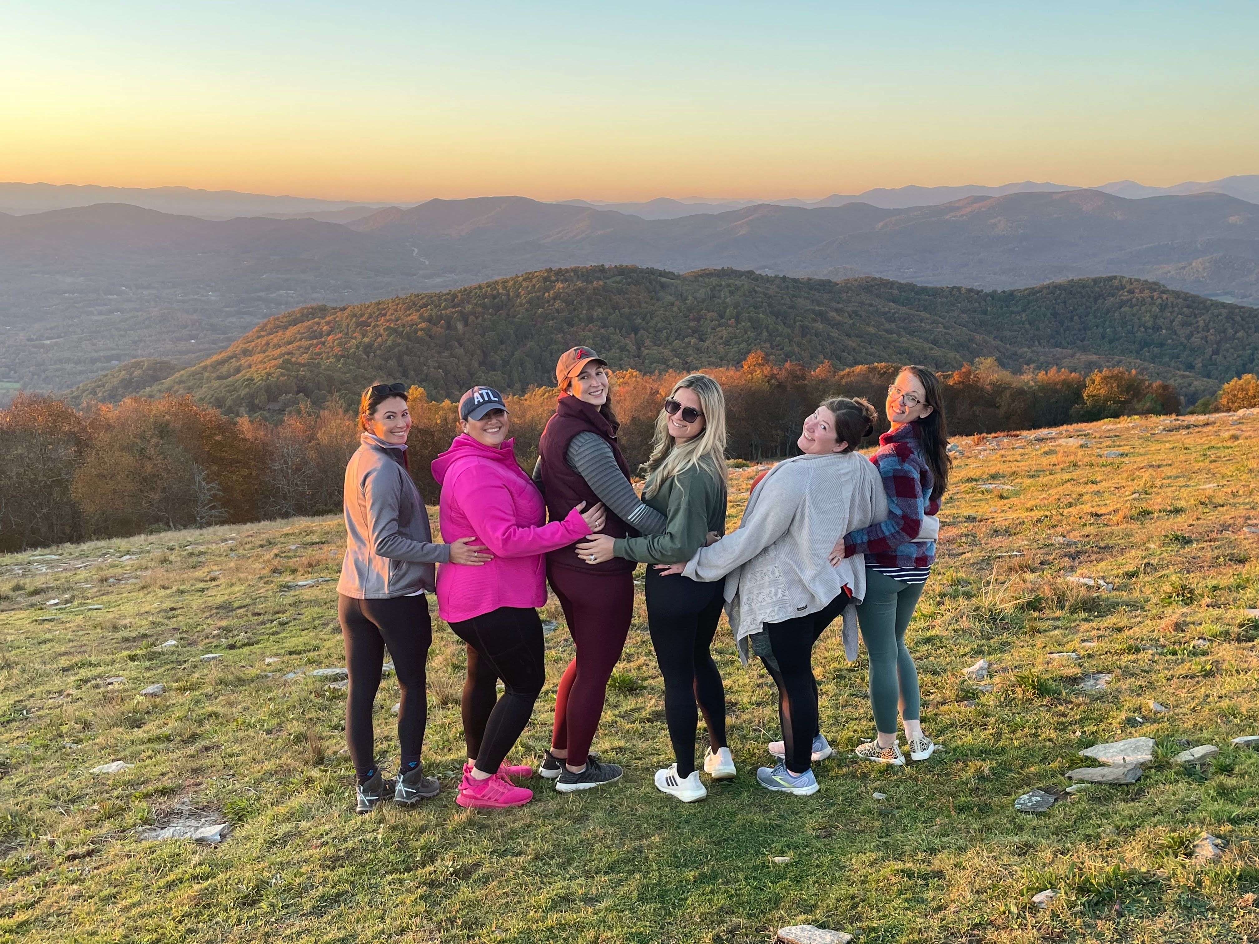 Six friends linking arms on a grassy mountain overlook at sunset, golden autumn trees and layered blue ridges in the distance.