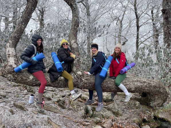 Four hikers in winter jackets with rolled yoga mats perched on a frost-covered tree trunk in a snowy mountain forest, smiling on a winter trail