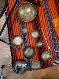 Assorted brass singing bowls and mallets arranged on a vibrant orange-red striped rug with blue geometric accents on a hardwood floor — meditation sound-healing set.