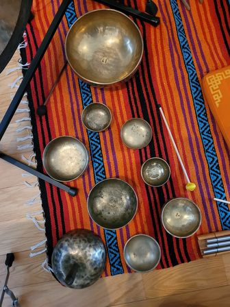 Assorted brass singing bowls and mallets arranged on a vibrant orange-red striped rug with blue geometric accents on a hardwood floor — meditation sound-healing set.