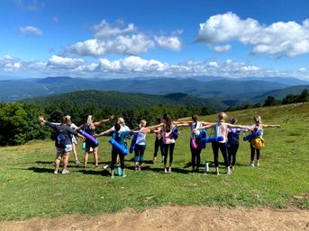 Group of hikers with rolled yoga mats standing on a grassy mountain ridge, arms outstretched toward a panoramic view of layered blue mountains and puffy clouds.
