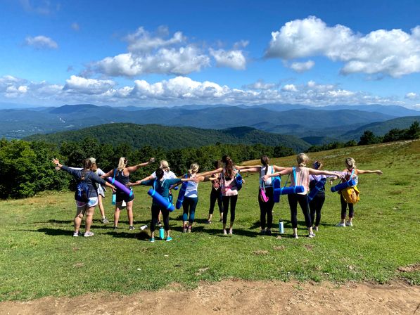 Group of hikers with rolled yoga mats standing on a grassy mountain ridge, arms outstretched toward a panoramic view of layered blue mountains and puffy clouds.