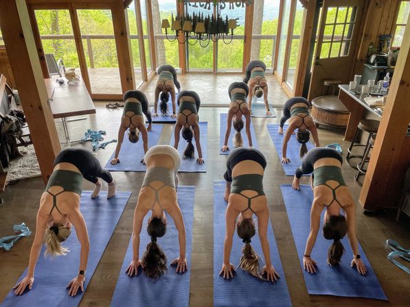 Group downward-dog yoga class in a rustic mountain cabin on blue mats, floor-to-ceiling windows framing lush green forest and distant hills, cozy retreat vibe.