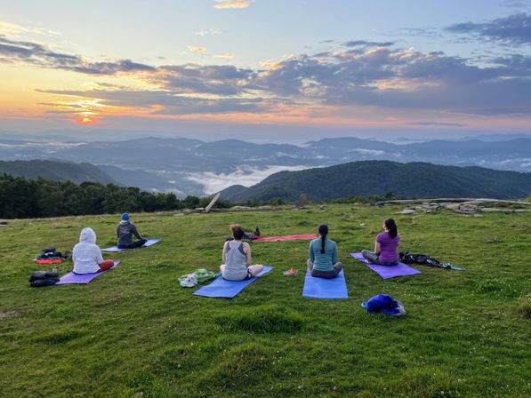 Five people seated on colorful yoga mats on a grassy mountaintop, watching a pink-orange sunrise over misty blue mountain ridges
