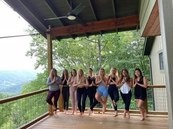 Outdoor yoga group on a covered wooden mountain deck practicing tree pose together, overlooking a lush forested valley with string lights overhead — yoga retreat vibe.