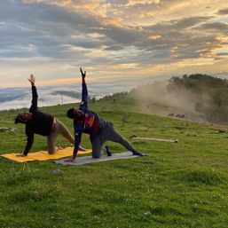 Two people practicing side-plank yoga on mats at sunrise atop a foggy mountain meadow with rolling hills and golden clouds