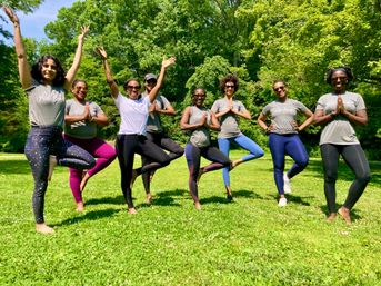 Eight people striking tree pose in a sunny green park — outdoor community yoga class in activewear and sunglasses, smiling and balancing on one leg.