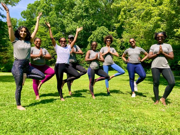 Eight people striking tree pose in a sunny green park — outdoor community yoga class in activewear and sunglasses, smiling and balancing on one leg.