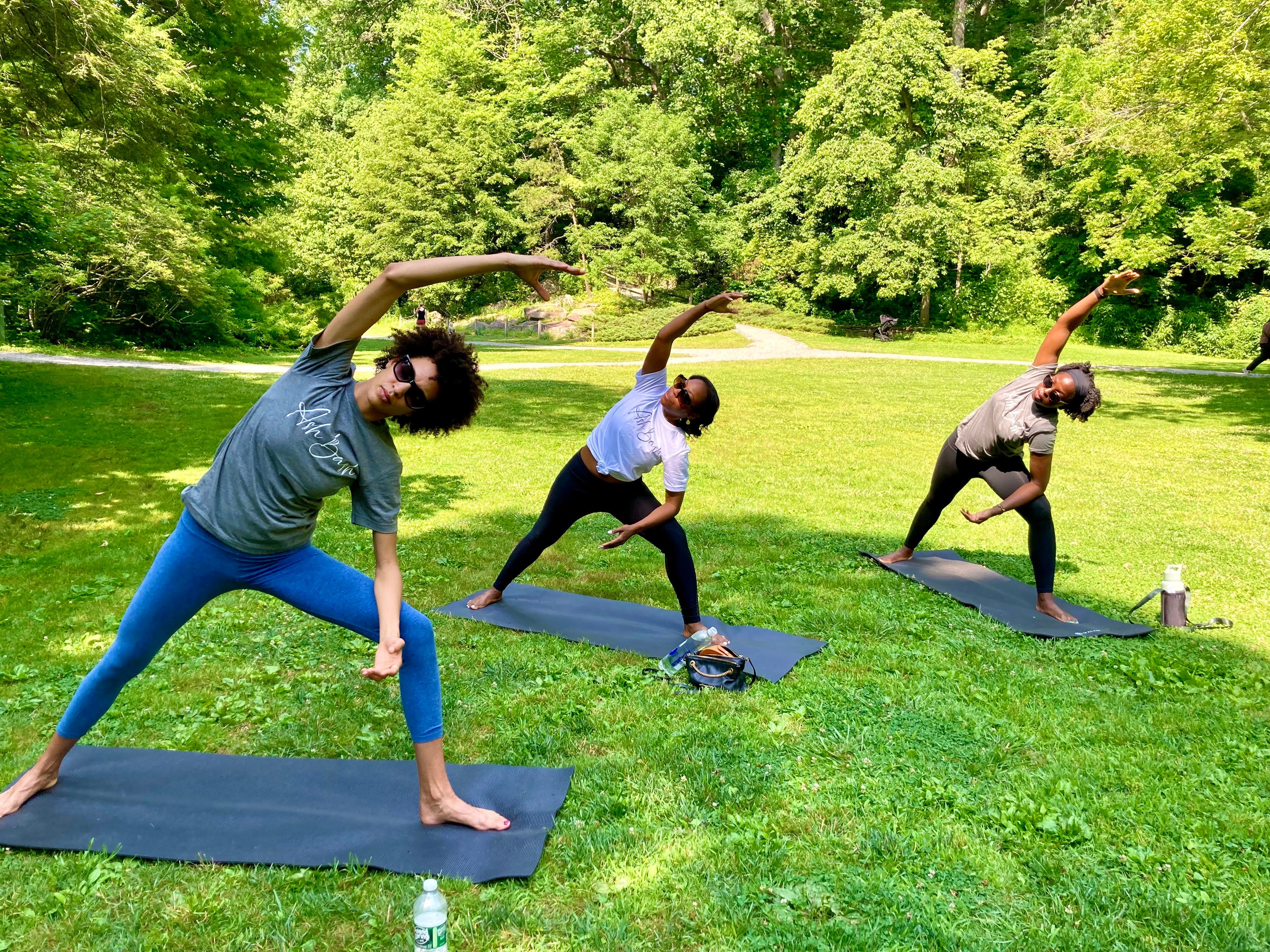 Three people on yoga mats doing side-stretch poses during an outdoor group yoga session in a sunny green park with trees in the background.
