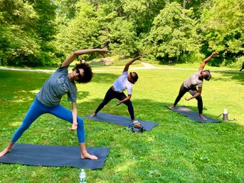 Three people on yoga mats doing side-stretch poses during an outdoor group yoga session in a sunny green park with trees in the background.