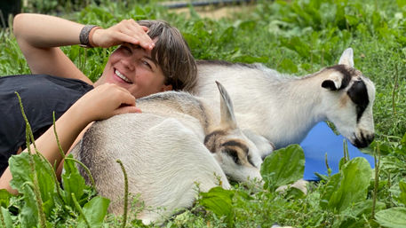 Smiling person lounging in a lush green pasture, cuddling two sleepy white-and-brown goats resting in the grass