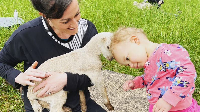 Toddler nuzzling a baby goat while an adult holds and pets it in a grassy field — playful family farm visit scene.