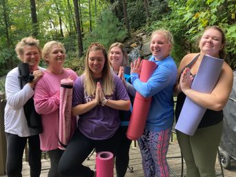 Six women holding rolled yoga mats and smiling with hands in prayer on a forested deck by a small waterfall — outdoor yoga retreat vibe.