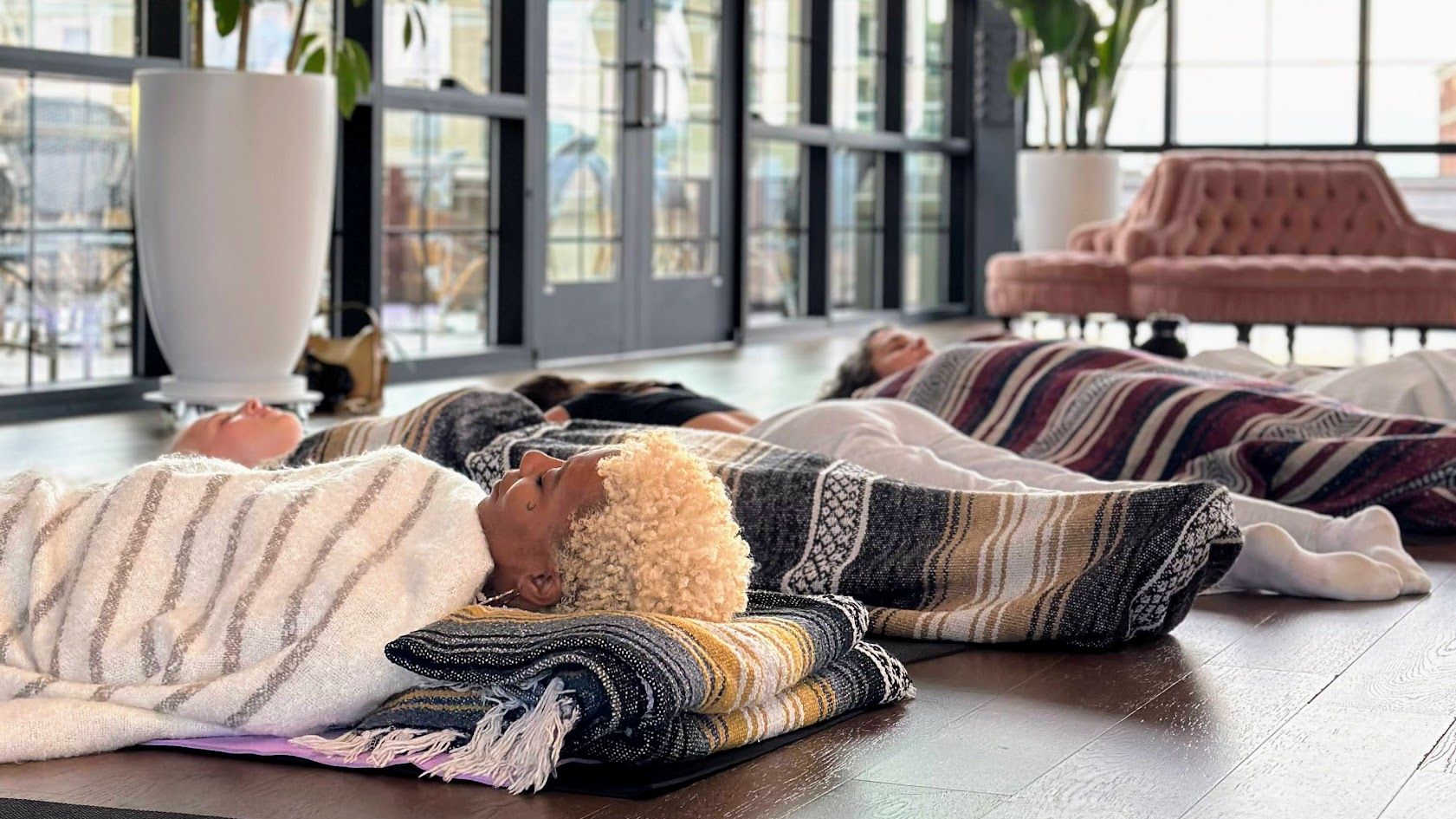 Cozy group relaxation in a sunlit wellness studio: people lying on a hardwood floor under striped woven blankets for restorative yoga/meditation, heads on folded blankets near large windows with plants and a tufted sofa in the background.