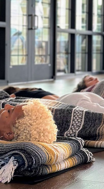 Cozy group relaxation in a sunlit wellness studio: people lying on a hardwood floor under striped woven blankets for restorative yoga/meditation, heads on folded blankets near large windows with plants and a tufted sofa in the background.