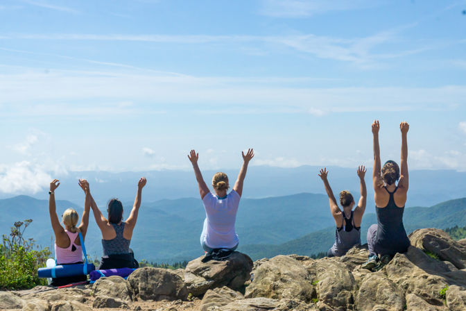 Five hikers cheer with arms raised on a rocky mountain overlook, looking out over layered blue ridges under a bright sky.