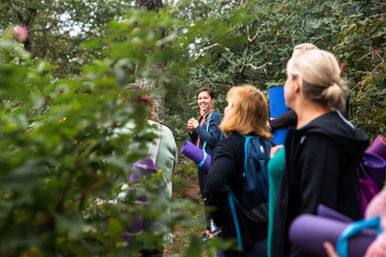 Smiling instructor leading a group of women carrying rolled purple yoga mats along a leafy woodland trail during an outdoor yoga retreat