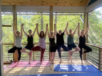 Seven people practicing yoga tree pose with arms raised and linked on a screened wooden deck overlooking lush green forested hills at a mountain retreat