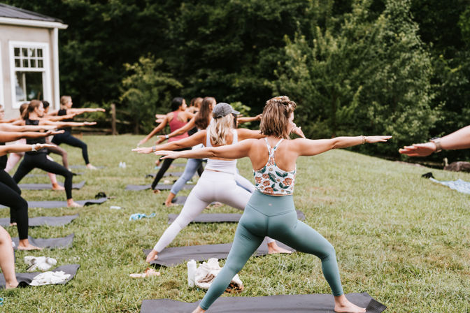 Outdoor yoga class participants practicing Warrior II on mats in a grassy park with lush trees, a community fitness and wellness session