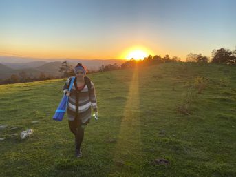 Person walking on a grassy hill at sunset carrying a blue yoga mat and water bottle, golden sun setting over rolling hills and trees in the background.