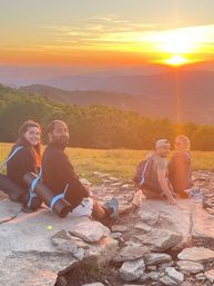 Group of four hikers with backpacks and rolled sleeping pads sitting on a rocky mountain overlook at golden sunset, layered ridges and forested valley below
