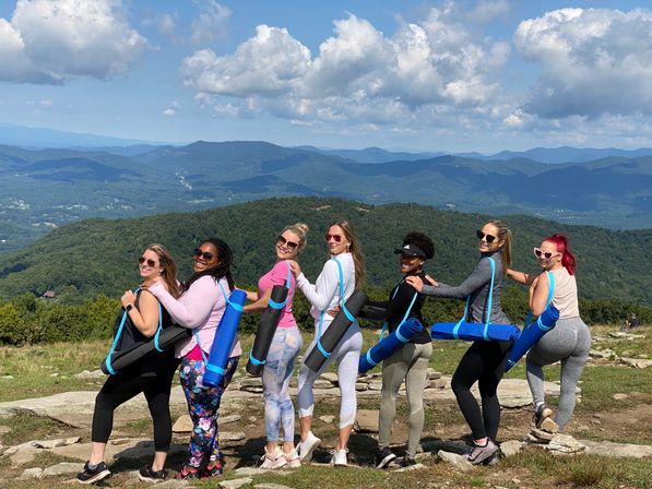Seven women in colorful activewear linked arm-in-arm carrying rolled blue yoga mats on a mountain overlook, smiling with layered blue ridges and puffy clouds in the scenic background