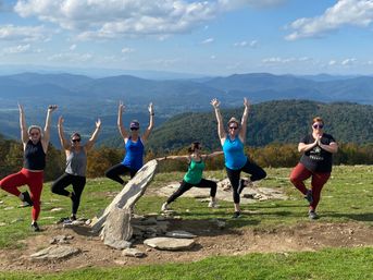 Six people doing yoga poses on a grassy mountaintop beside a stone cairn, sunlit and playful, with sweeping layered blue mountain ridges and a partly cloudy sky.