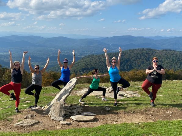 Six people doing yoga poses on a grassy mountaintop beside a stone cairn, sunlit and playful, with sweeping layered blue mountain ridges and a partly cloudy sky.