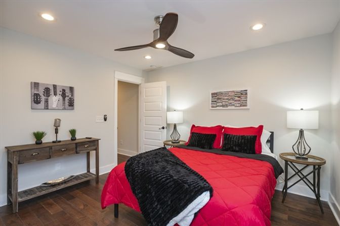 Contemporary bedroom with bold red comforter and black throw on a bed, matching red and black pillows, hardwood floors, wooden console table, round bedside tables with lamps, ceiling fan, light-gray walls and framed wall art.