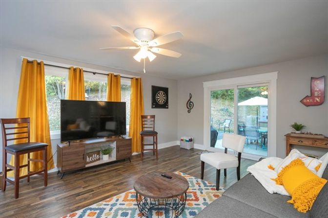 Cozy contemporary living room with mustard-yellow curtains, flat-screen TV on a mid-century console, patterned area rug, gray sofa with yellow pillows, ceiling fan, hardwood floors and sliding glass door to a backyard patio.