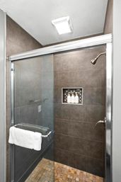 Sleek modern glass-enclosed shower with sliding door, dark gray wall tiles, mosaic stone floor, recessed niche holding three bottles, chrome showerhead and towel bar with white towel.