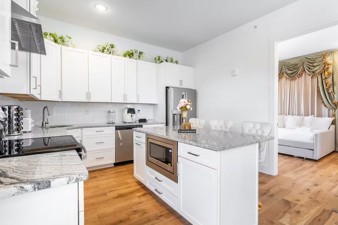 Bright, stylish modern white kitchen with marble-look countertops, central island with built-in microwave and bar stools, stainless appliances, hardwood floors, greenery above cabinets, and a view into an adjacent living room with a tufted sofa and ornate drapery.