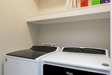 Bright compact laundry nook with matching white washer (top-load) and dryer (front-load) under a white shelf holding detergent and supplies — clean, modern laundry room.