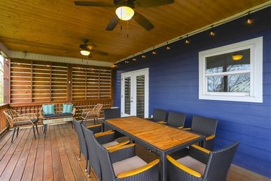 Covered wooden deck with blue exterior siding, large wooden dining table with wicker chairs for eight, ceiling fans, string lights and a cozy rattan lounge area.
