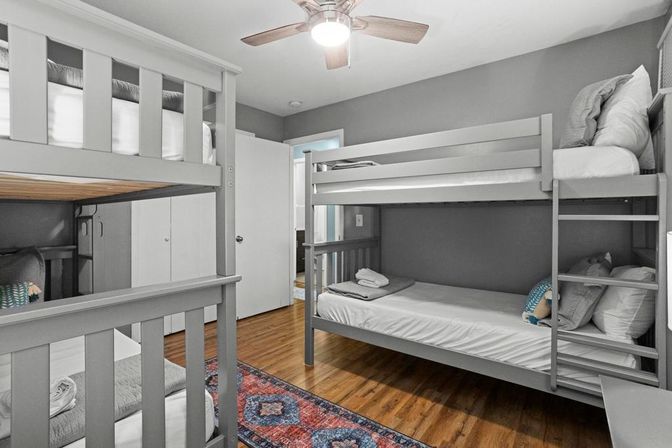 Cozy shared bedroom with gray wooden bunk beds, hardwood floors, colorful patterned rug, ceiling fan and neatly made beds with folded towels.