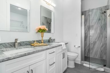 Bright modern white bathroom with double-sink vanity, gray granite countertop, gold vase of pink roses, two mirrors, glass-enclosed marble-tiled shower and toilet.