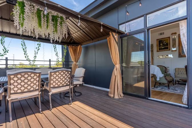Rooftop patio with a covered pergola draped in hanging white wisteria and string lights, outdoor dining table with cushioned lattice-back chairs on a wood deck, tan curtains tied back and a sliding glass door opening into a cozy living room.