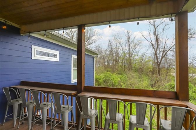 Covered wooden balcony with bar-height counter and a row of metal stools, blue house siding and string lights overhead, overlooking a green wooded backyard.