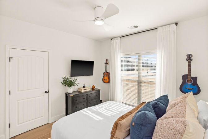 Sunlit modern bedroom with white walls, cozy bed with blue and beige pillows, black dresser and wall-mounted TV, two acoustic guitars hanging on the wall, ceiling fan and large window with white curtains.